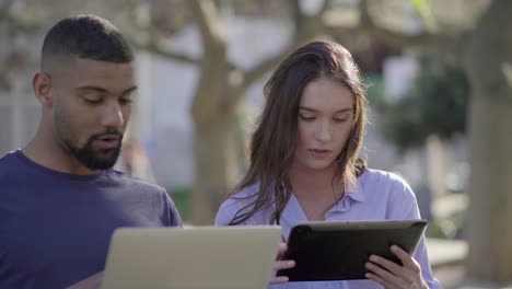 Man-and-woman-in-park-discussing-something-on-laptop-and-tablet